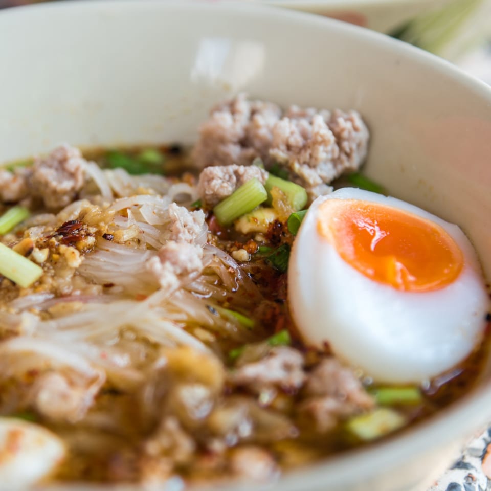 Close-up of Thai noodle soup with minced pork, green onions, and a soft-boiled egg.