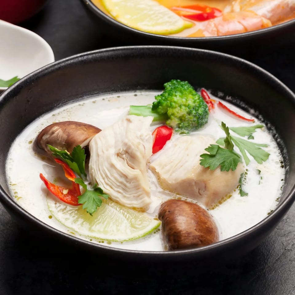 Close-up of Tom Kha Gai soup with chicken, mushrooms, broccoli, chili, and lime in a black bowl.